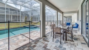Uma área externa coberta com piscina, mesa de jantar com cadeiras e vista para uma casa através de um compartimento de vidro.
