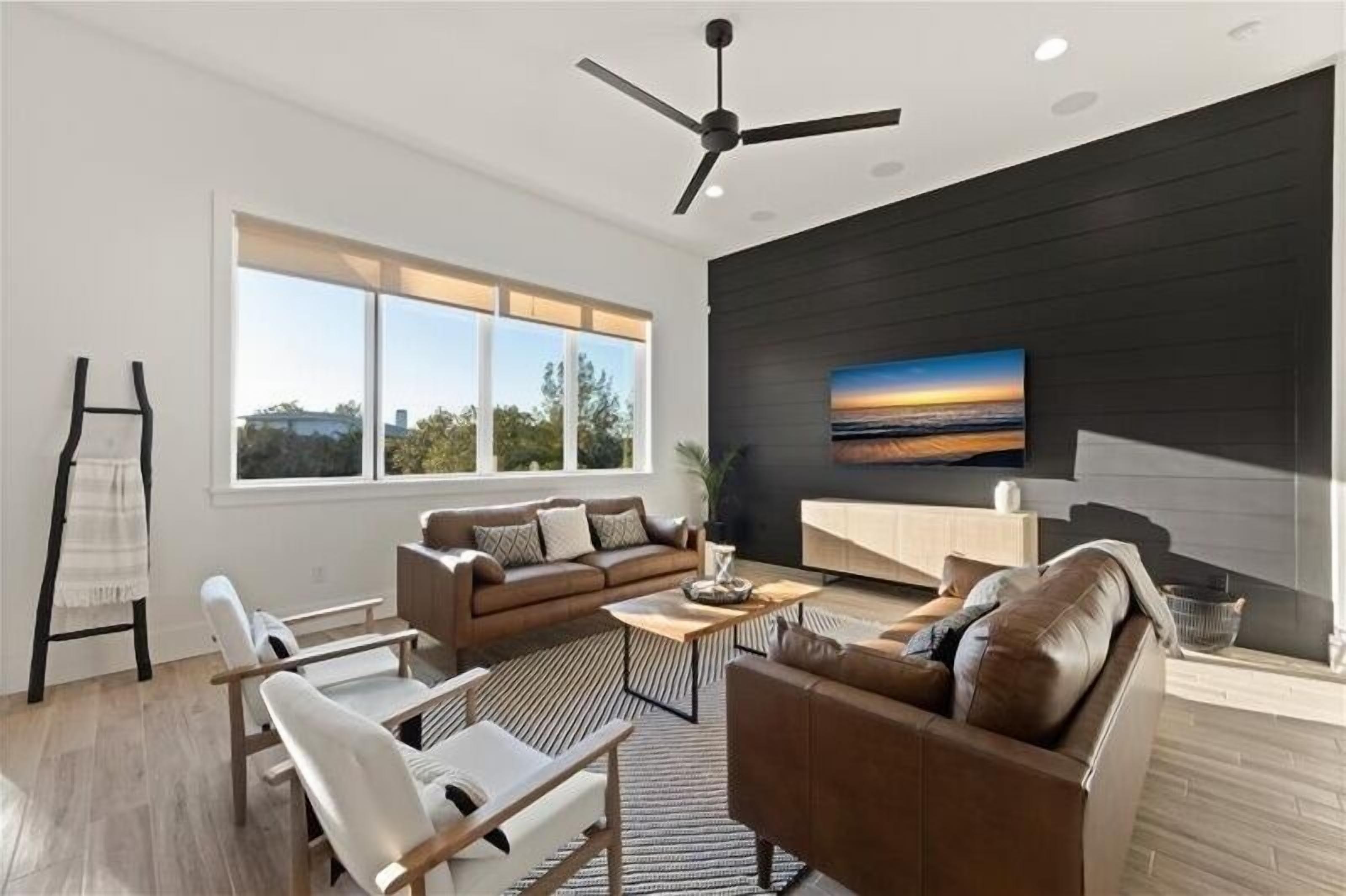 A modern living room with a large window, a ceiling fan, and a flat-screen TV mounted on the wall.