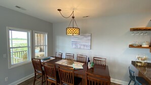 A dining area with a wooden table and chairs, a hanging light fixture, and a view through large windows. - Waterfront: Dock Access, Walk to Beach, Pier & Shops, 5BR, Sunset view / 10% off (Surf City)