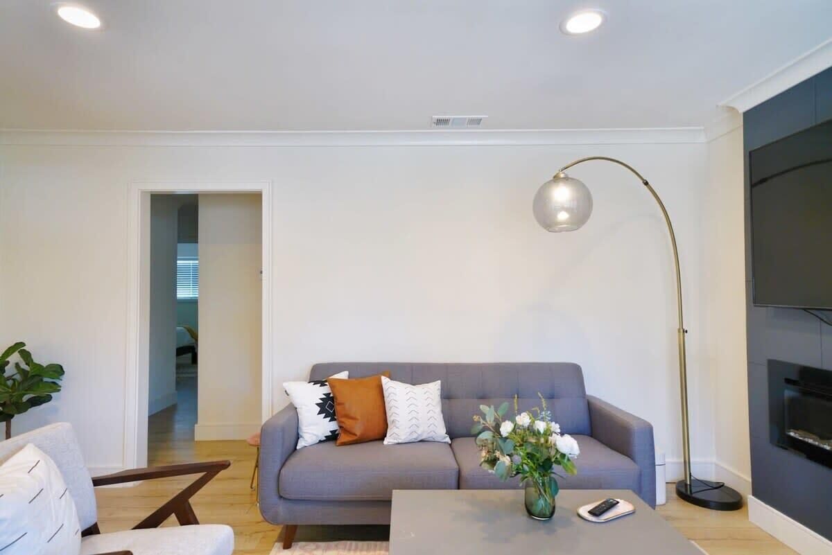 Una sala moderna con un sofá gris, una mesa de centro con un florero, una lámpara de pie y un televisor de pantalla plana montado en la pared.