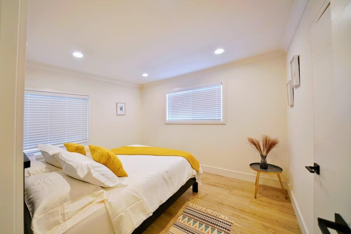 A bedroom with a bed, two windows with blinds, a small table with a vase, and a door.