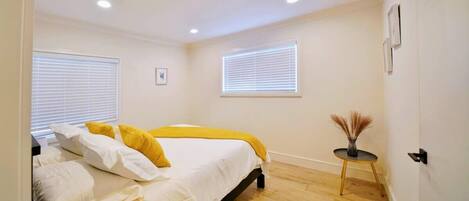 A bedroom with a bed, two windows with blinds, a small table with a vase, and a door.