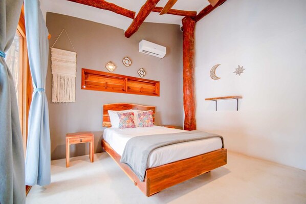 Une chambre avec un lit en bois, une table de chevet, une décoration murale en macramé suspendue, un climatiseur fixé au mur et une étagère en bois ornée d’un croissant de lune et d’une étoile.