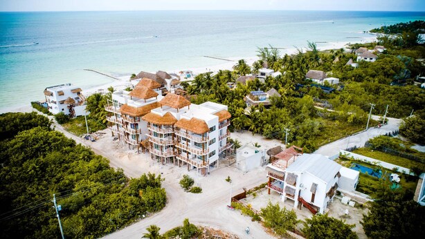 Een resort met meerdere gebouwen, waaronder een groot centraal gebouw met meerdere balkons en rieten daken, omgeven door weelderige begroeiing en een strand.