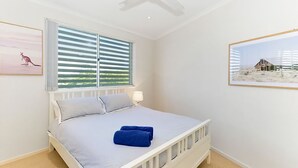 A bedroom with a bed, a window with blinds, a lamp, and two framed pictures on the walls. - With stunning ocean views and open living, enjoy the coastal lifestyle of Beauty on Bowra. (Nambucca Heads)