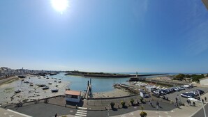 A coastal marina with boats, a small building, and a paved area with cars and people.