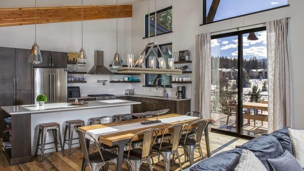 A modern kitchen with a central island, dining area with chairs, and a large window with a view of a snowy landscape.