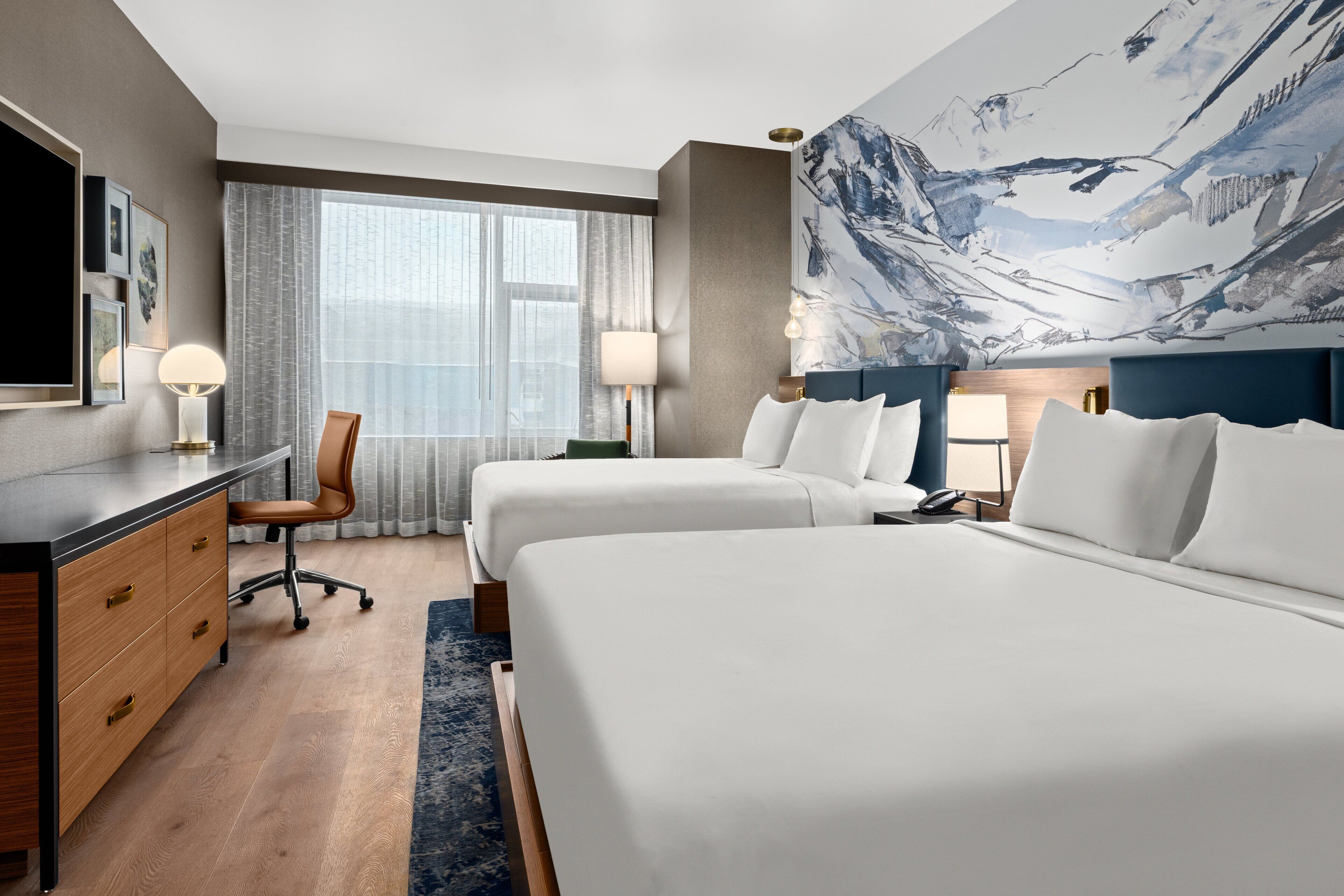 Ett hotellrum med två sängar, ett skrivbord, en stol och ett stort fönster med gardiner.