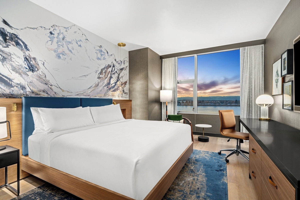 Ein Hotelzimmer mit einem großen Bett, einem Schreibtisch, einem Stuhl und Blick auf die Stadtlandschaft durch das Fenster.