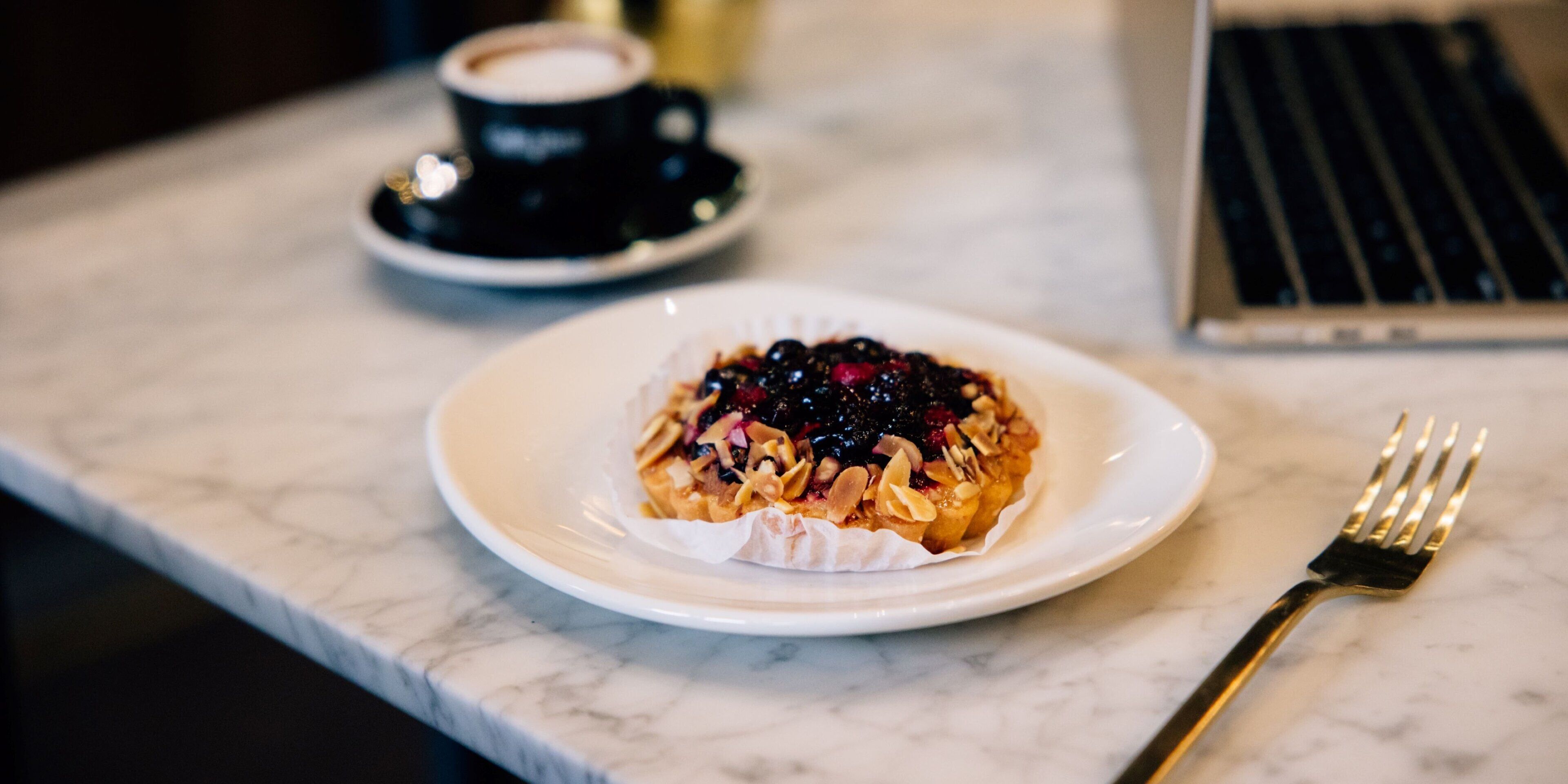 A dessert on a plate with a fork beside it, a cup of coffee, and a laptop on a marble surface.