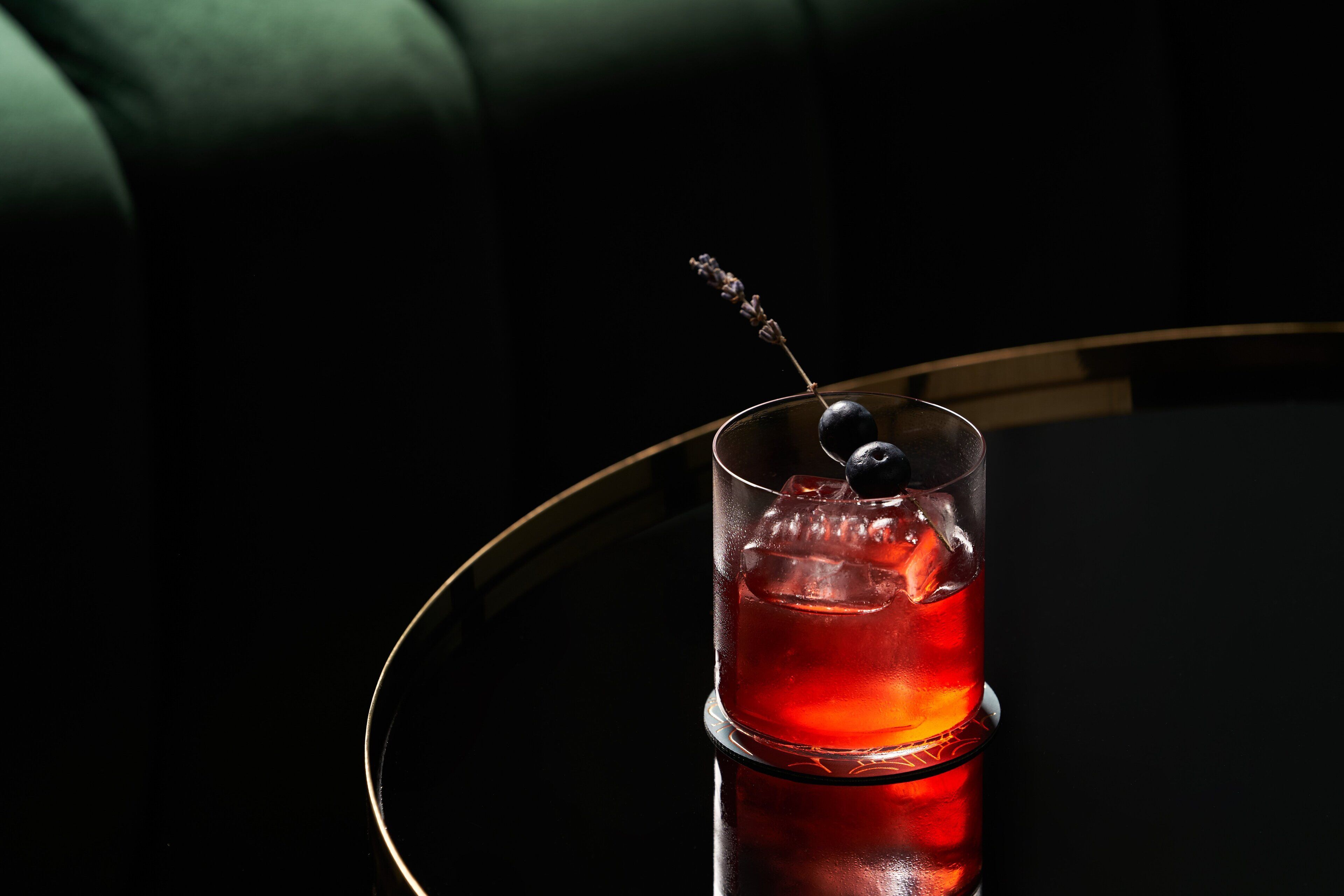 Un bicchiere con una bevanda rossa e un'oliva decorativa su una superficie riflettente.
