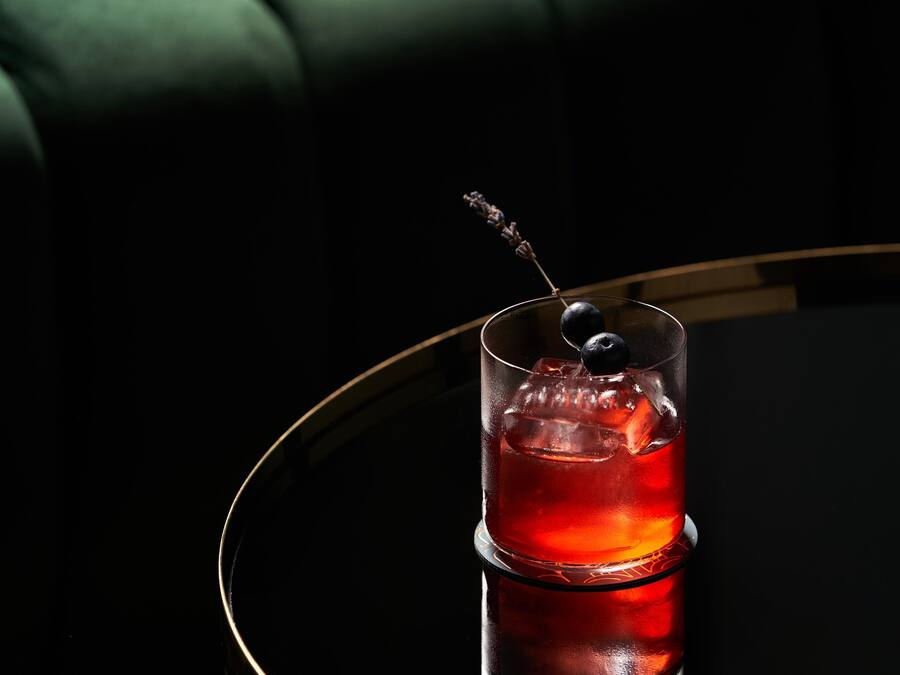 Un bicchiere con una bevanda rossa e un'oliva decorativa su una superficie riflettente.
