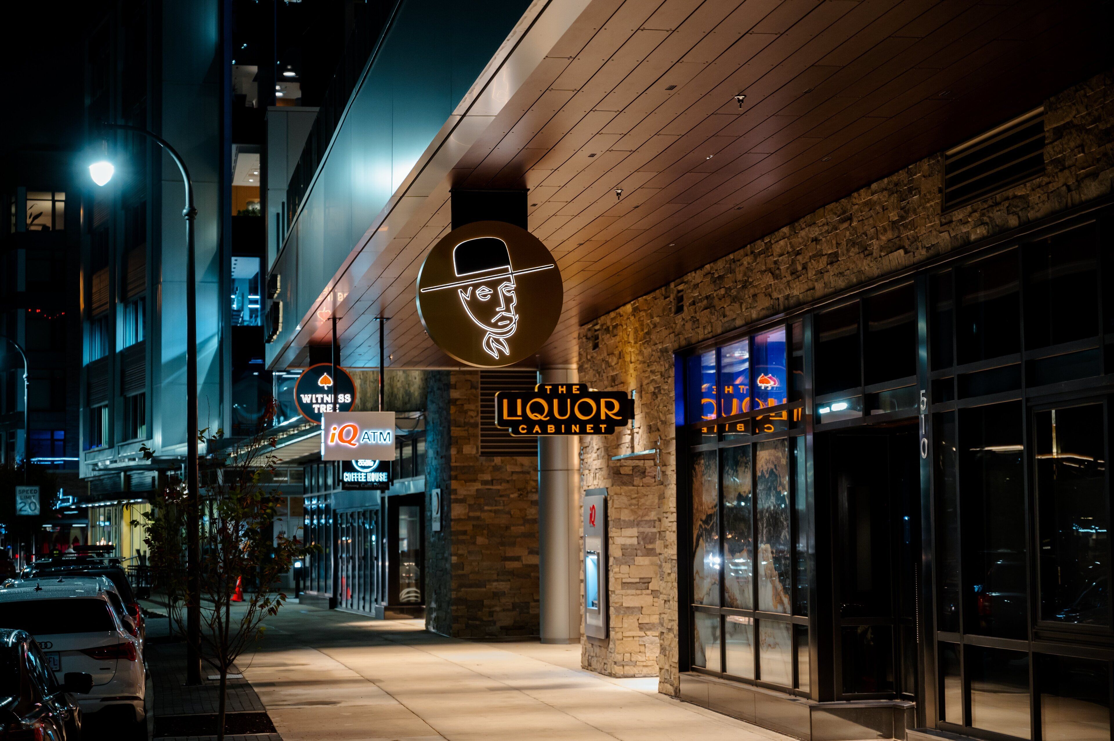 A nighttime urban street with a building featuring a neon sign for 'The Liquor Cabinet' and a 'With Us' sign.