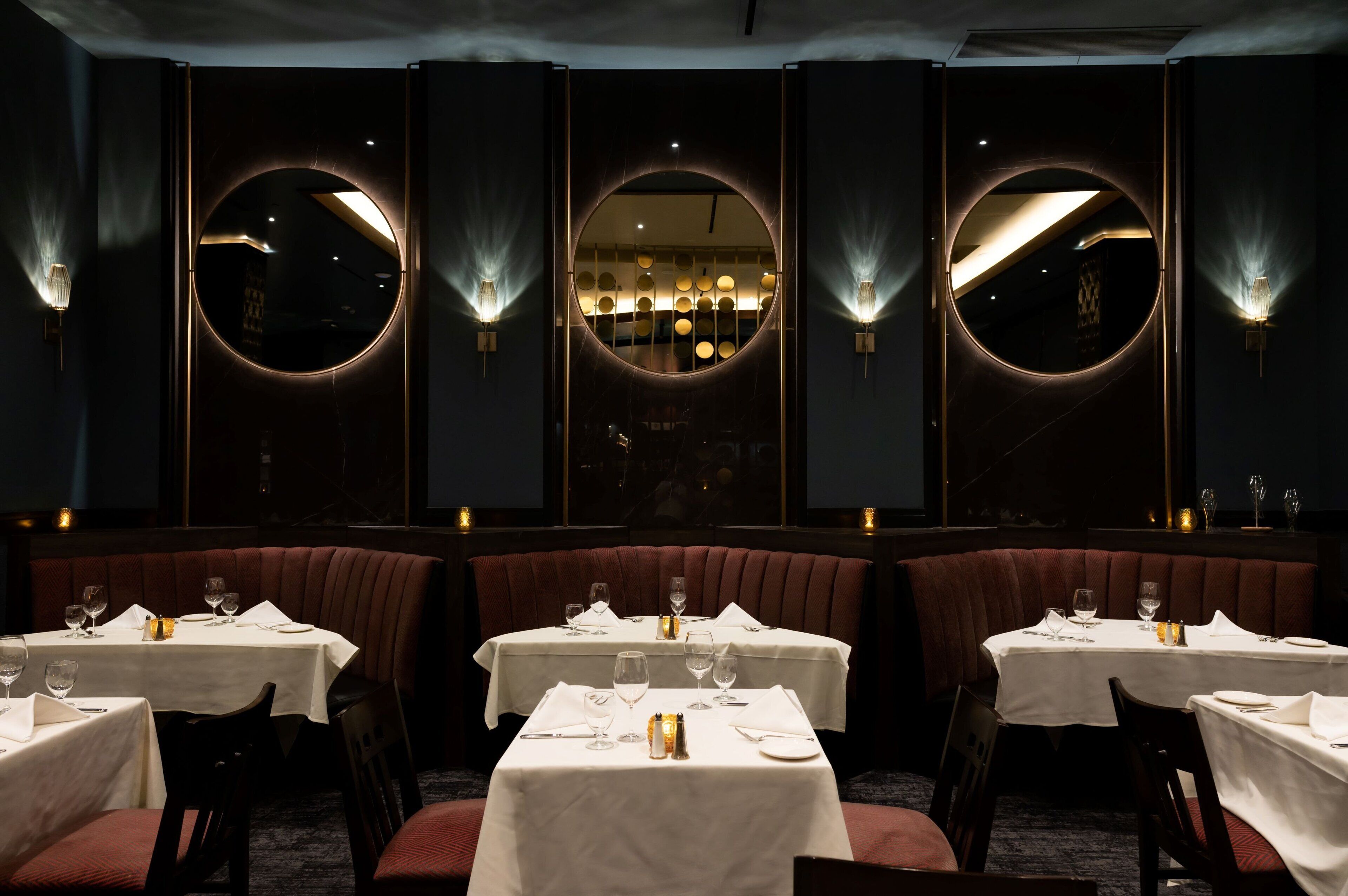 En restaurang med runda speglar och vägglampor, borden är dukade med vita dukar och glas.