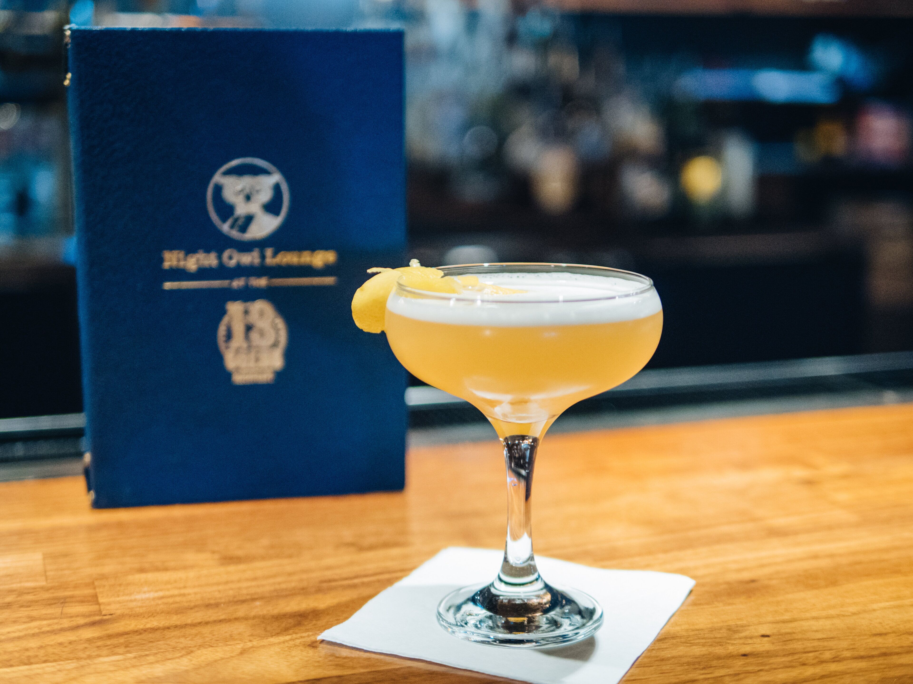 A cocktail in a stemmed glass with a lemon garnish on a wooden table. In the background, there is a blue box with a logo and text.
