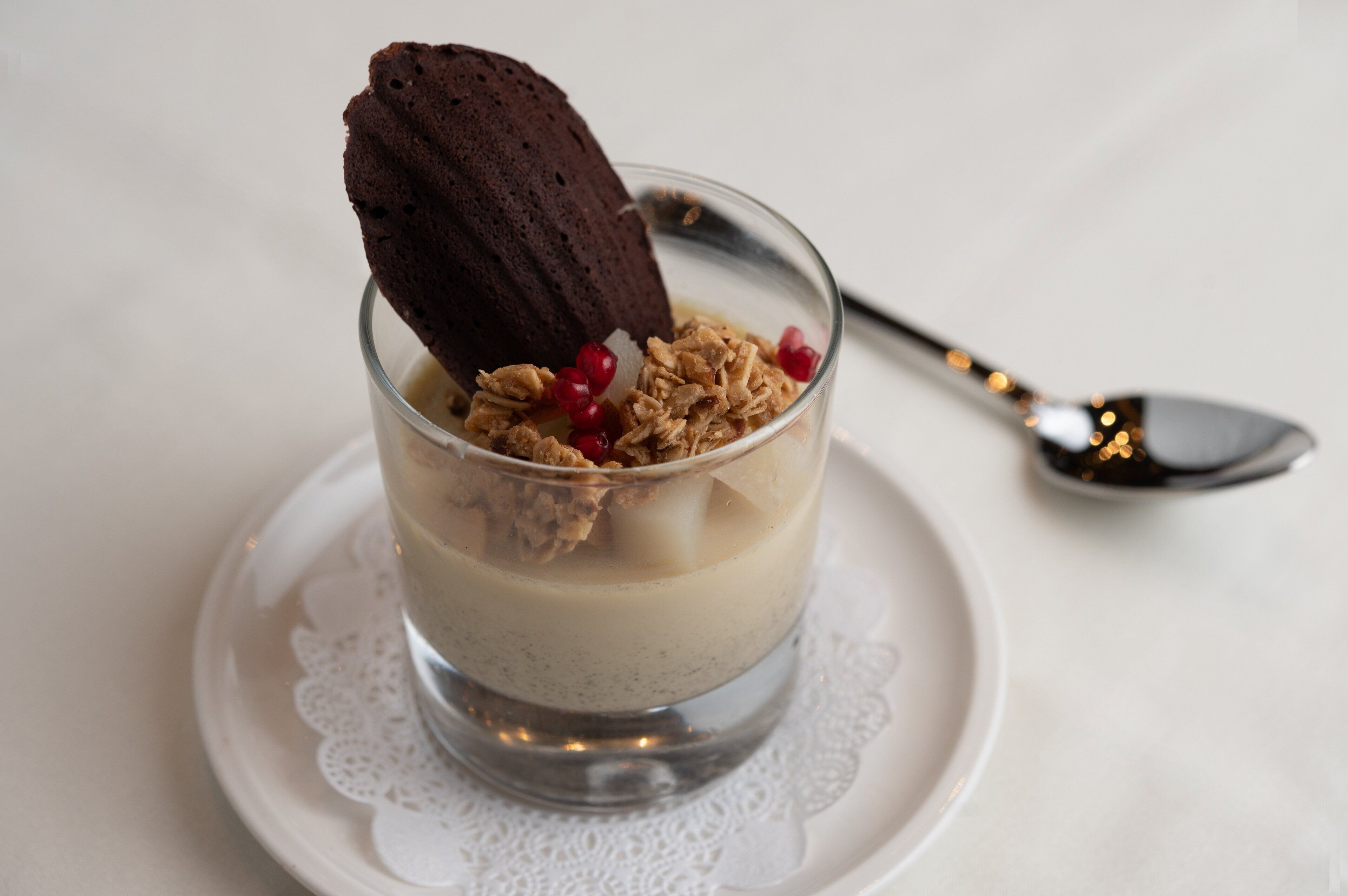 Un dessert servi dans un verre transparent, garni d’un biscuit et de granola, présenté sur une assiette blanche avec une cuillère à côté.