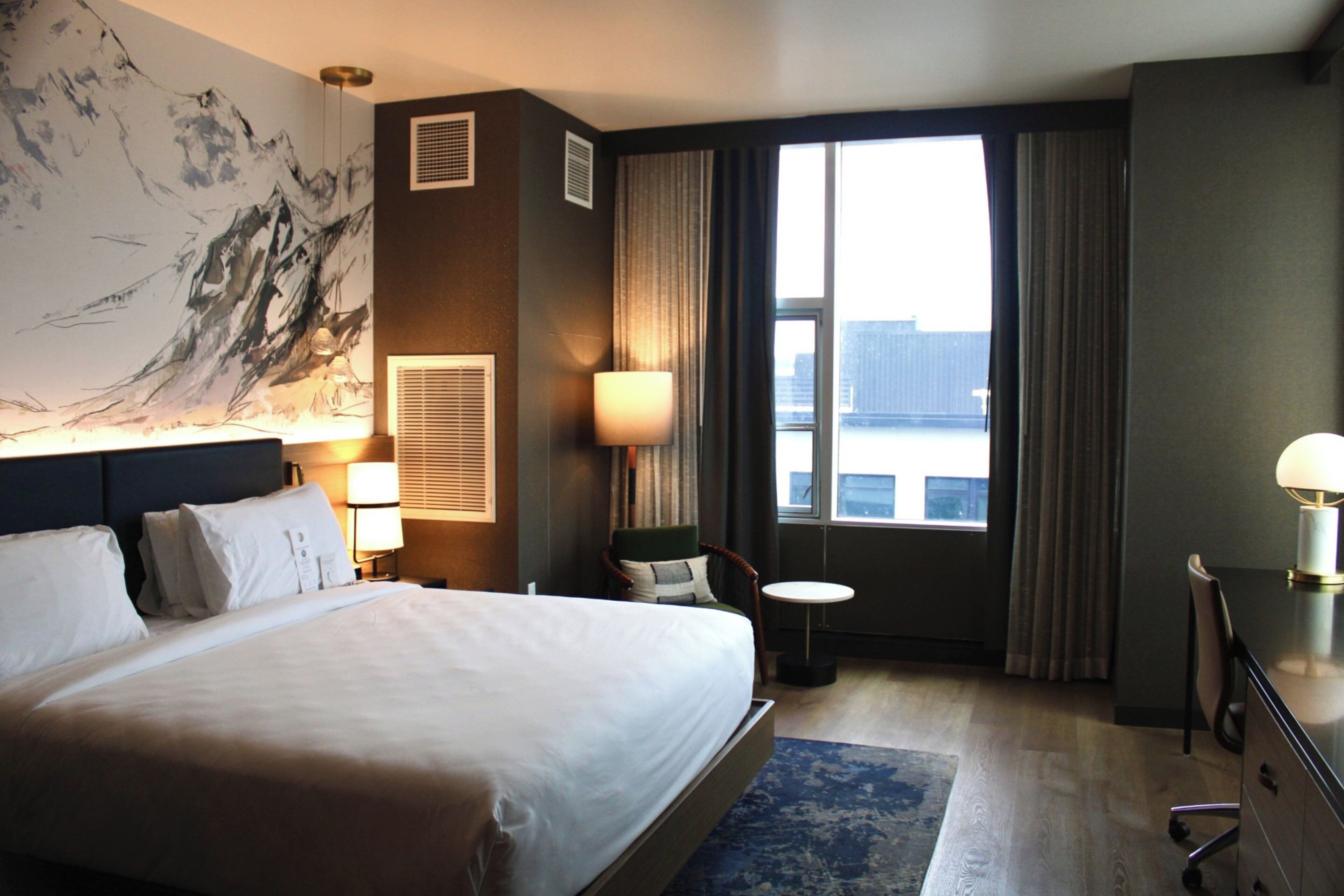 Ein Hotelzimmer mit einem großen Bett, einem Schreibtisch, einem Stuhl, einem kleinen Tisch und einem großen Fenster mit Vorhängen.