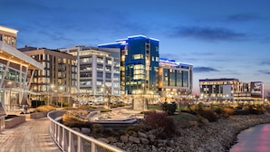 Een modern complex van stadsgebouwen met meerdere hoge torens, een aangelegde zone met zitplaatsen en een rivier op de voorgrond.