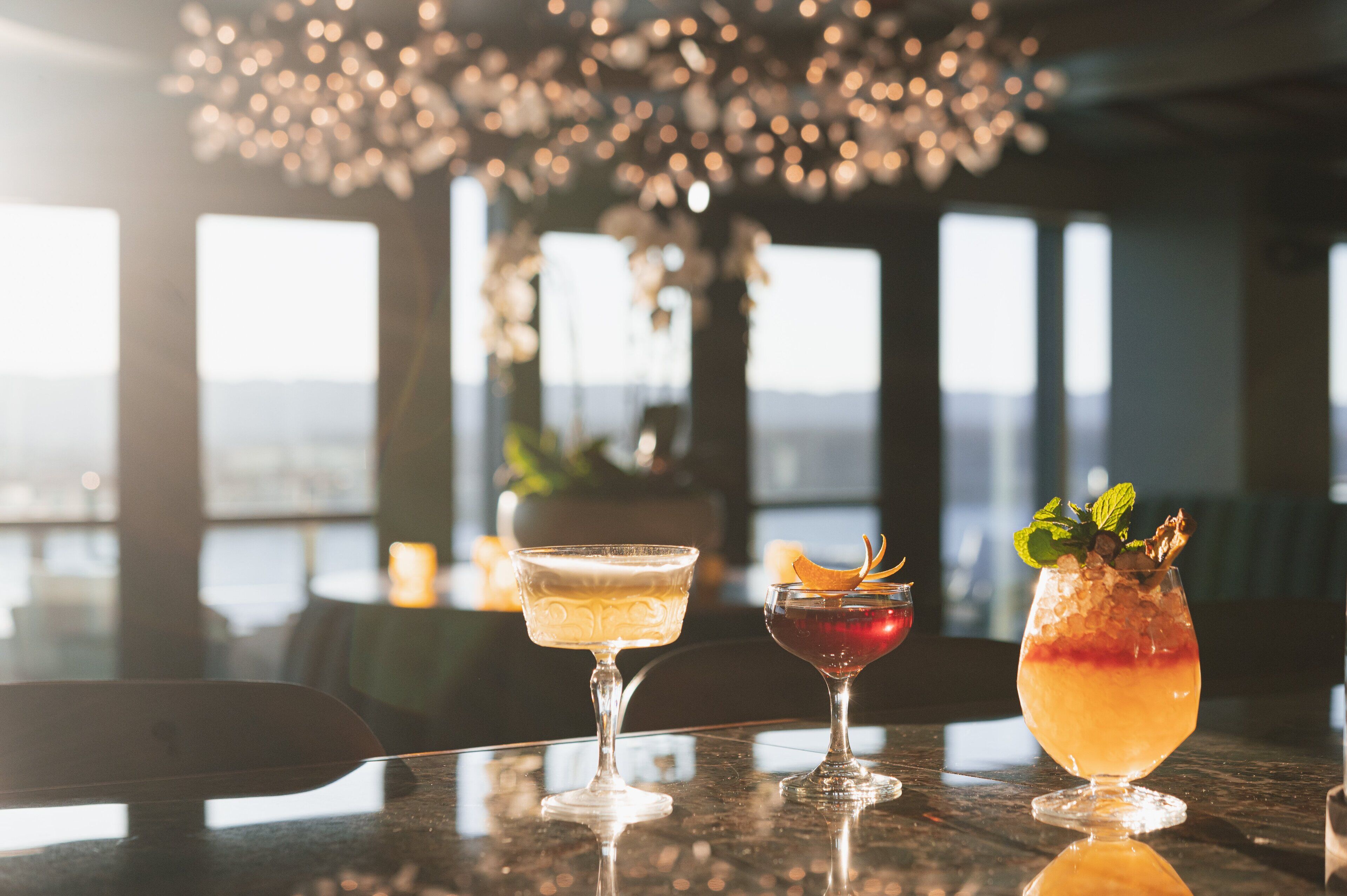 Three cocktails on a bar counter with a view of the cityscape through large windows.