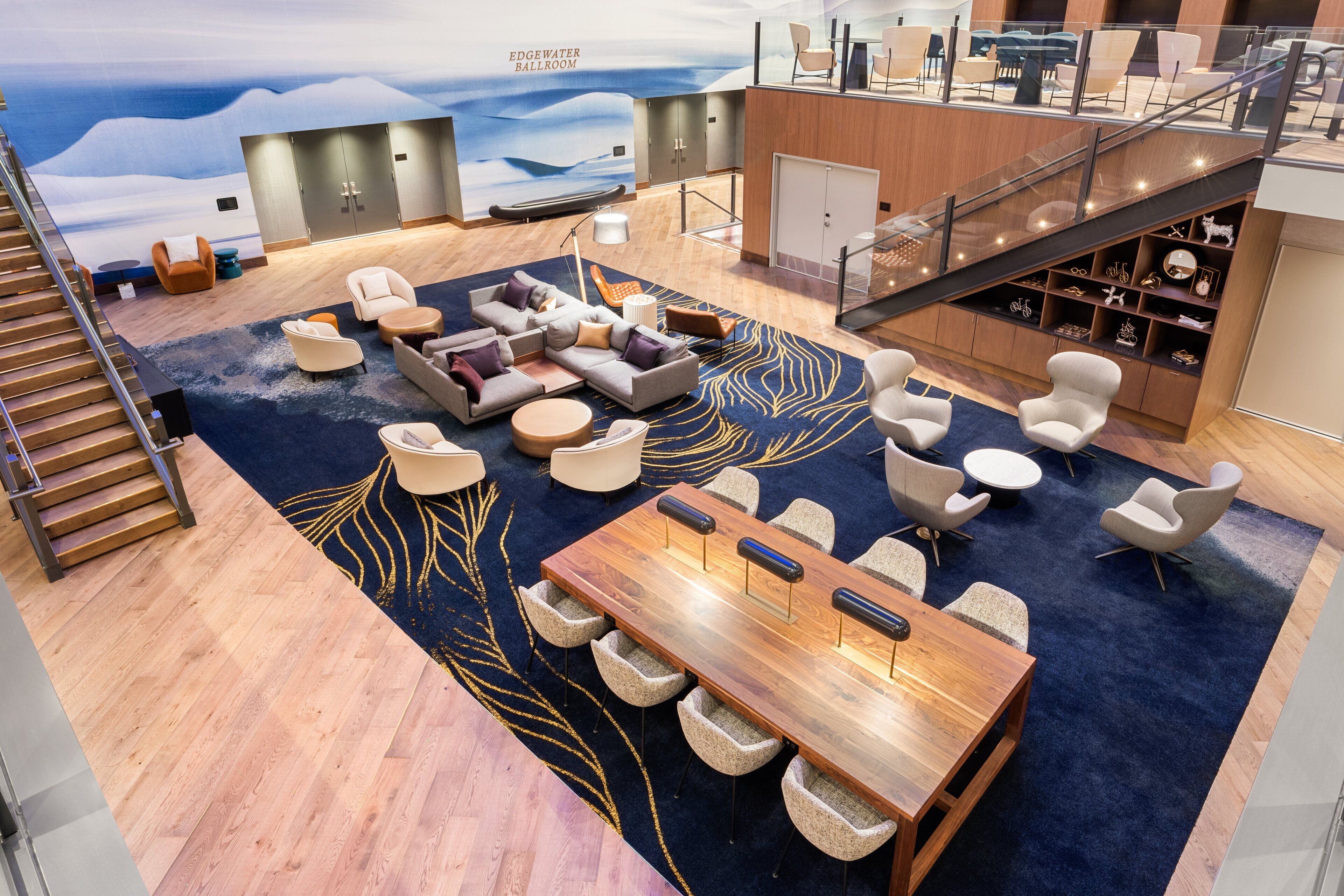 Un área de estar moderna con una gran mesa de madera, sofás y sillones, una escalera y vistas al salón de baile Edgewater.