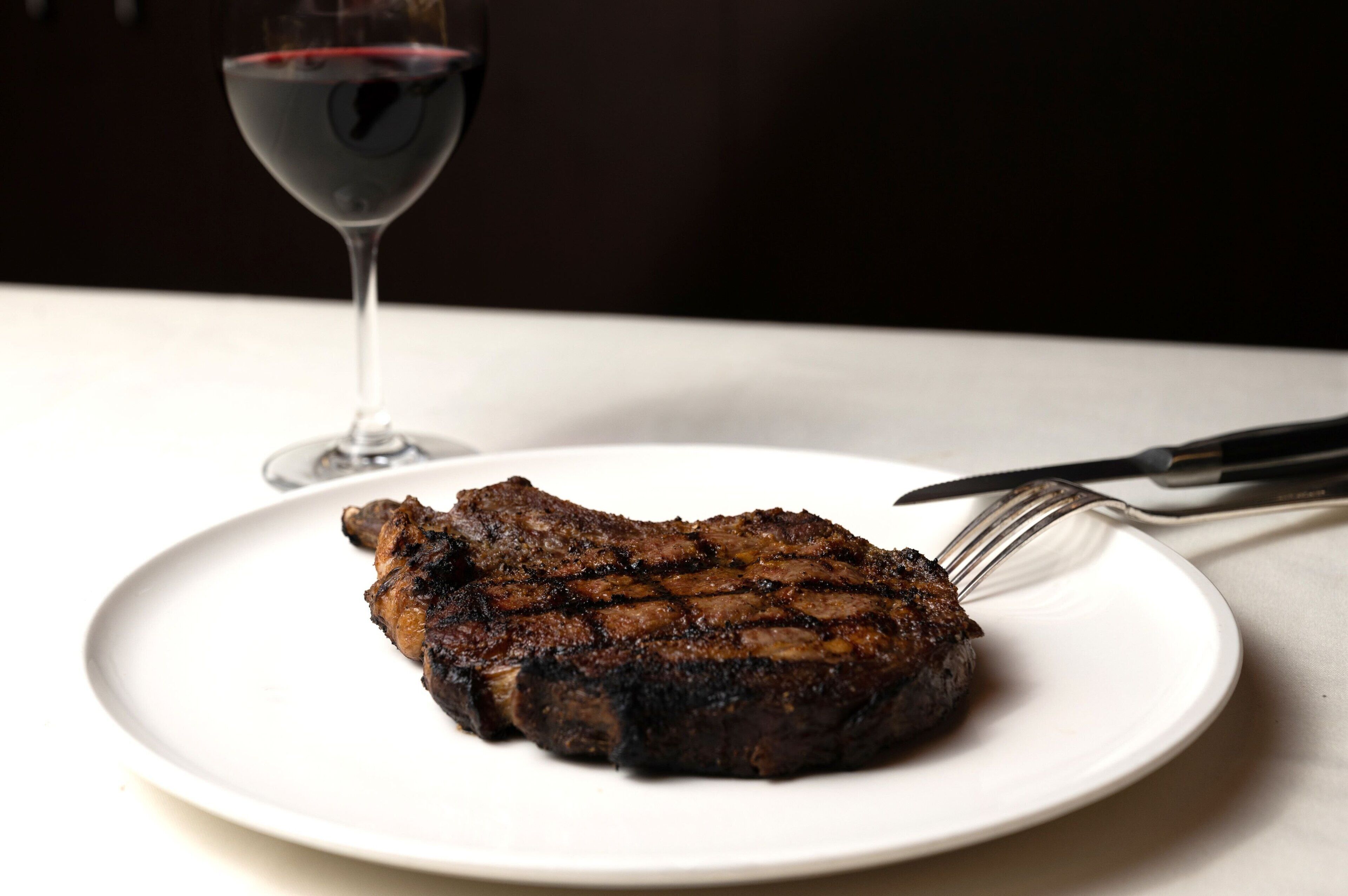 Un steak sur une assiette blanche, avec une fourchette et un couteau à côté, et un verre de vin rouge en arrière-plan.