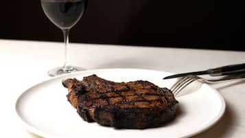 Un steak sur une assiette blanche, avec une fourchette et un couteau à côté, et un verre de vin rouge en arrière-plan.