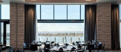 A dining area with tables set for a meal, large windows with a view of a body of water and distant landscape, and modern chandeliers.