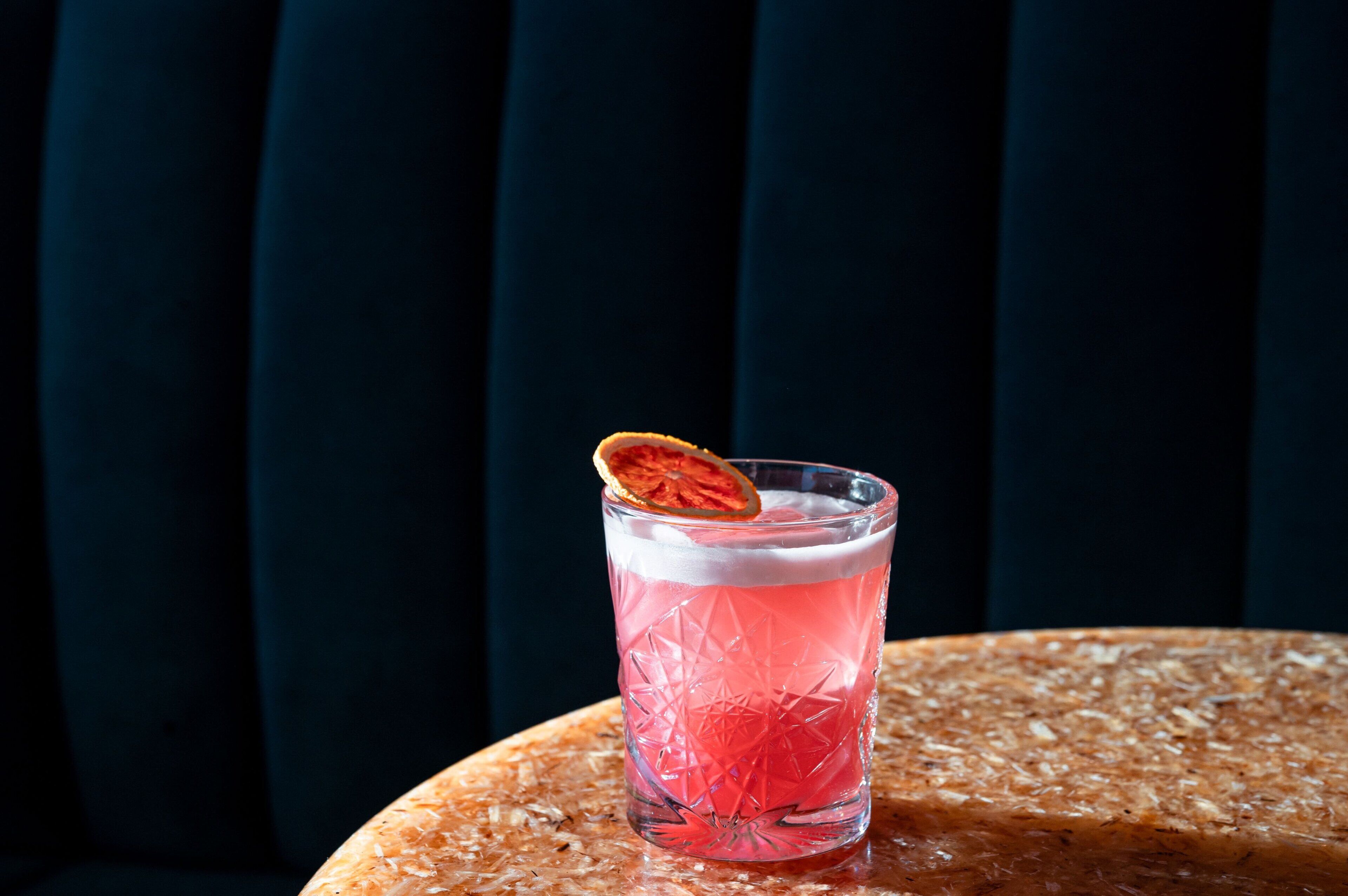 Un vaso de bebida rosada con una guarnición de fruta deshidratada sobre una superficie texturizada.