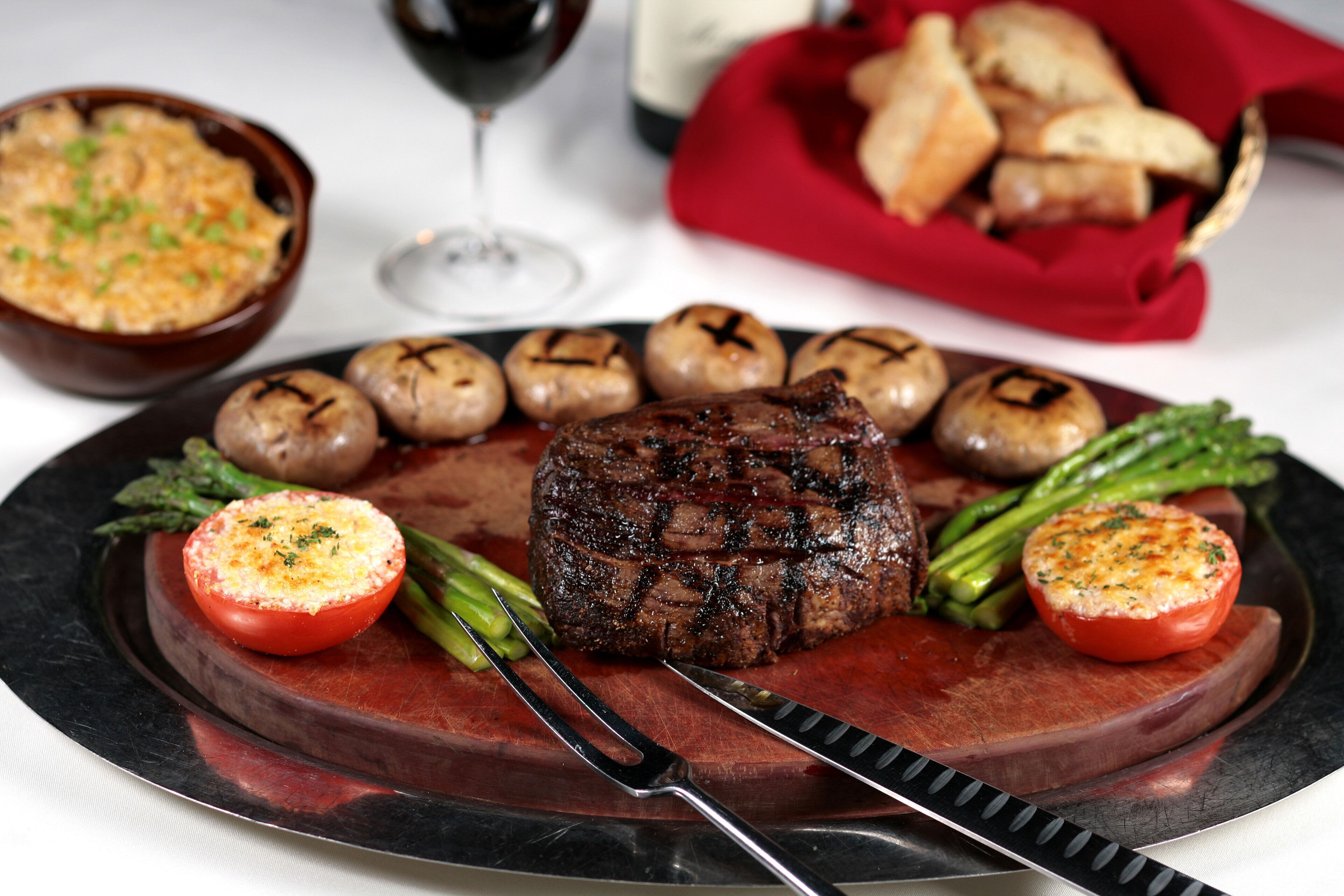 Een steak met gegrilde champignons en asperges op een bord, vergezeld van een soepkom en een glas rode wijn.