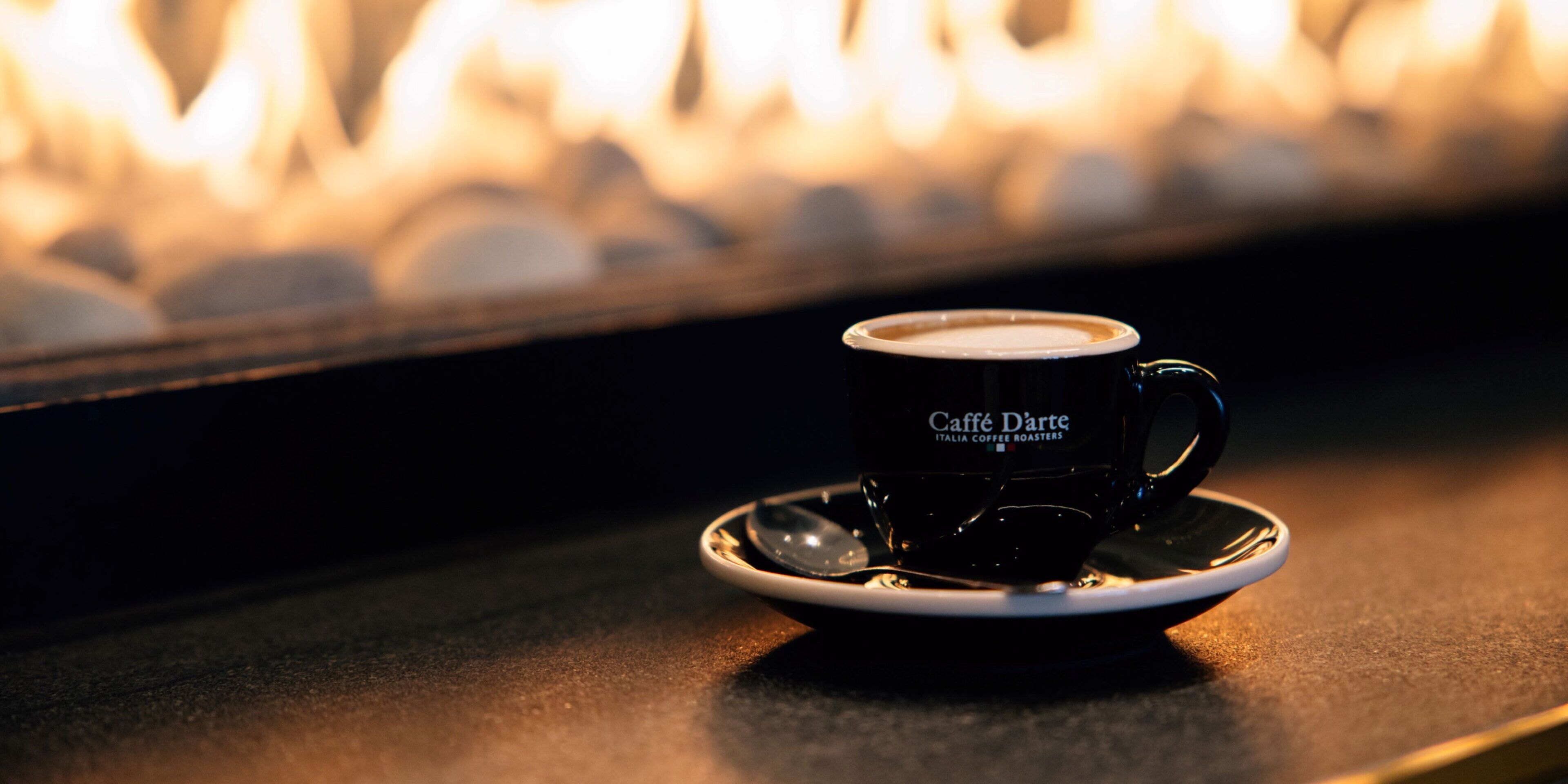 A cup of coffee on a saucer with a blurred background of a fireplace.