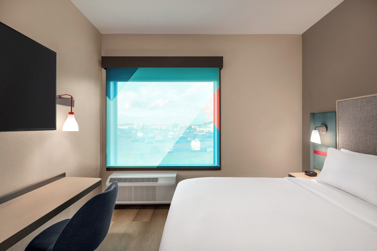 Hotellihuone, jossa on sänky, työpöytä, tuoli, ikkunasta avautuva näkymä ja seinään kiinnitetty valaisin.