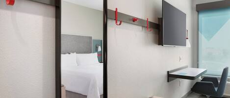A hotel room with a bed, a desk, a chair, a TV mounted on the wall, and a window.