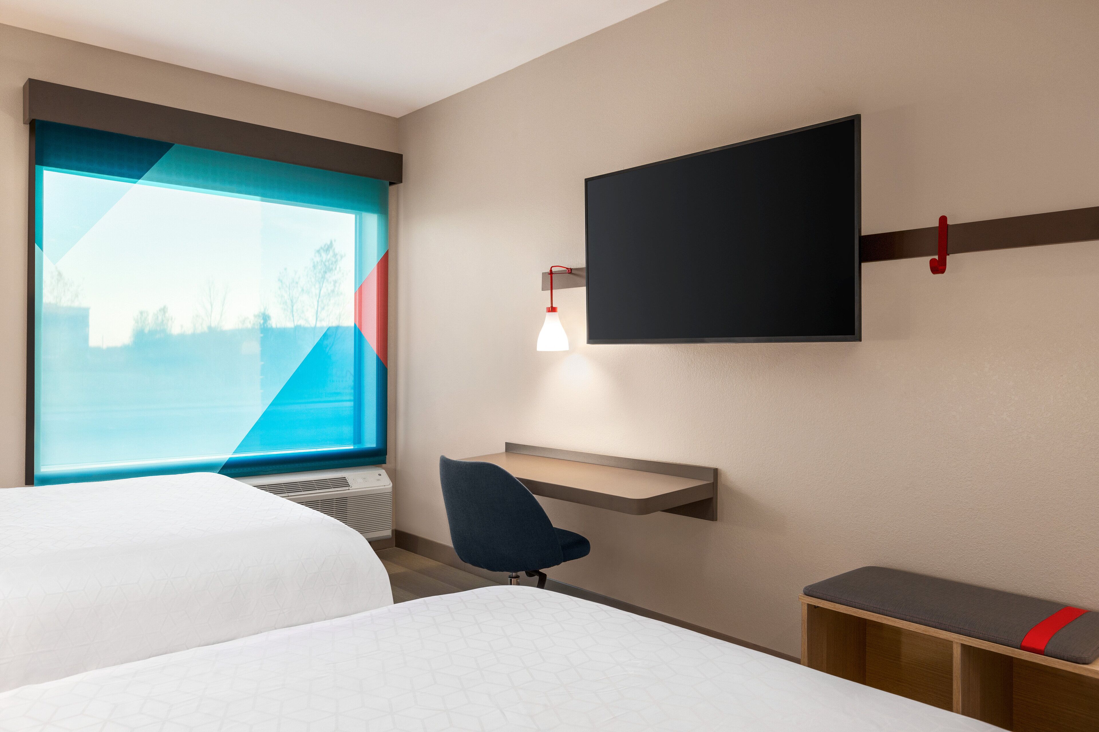 Habitación de hotel con dos camas, un escritorio, una silla, un televisor montado en la pared y una ventana con vistas a árboles.