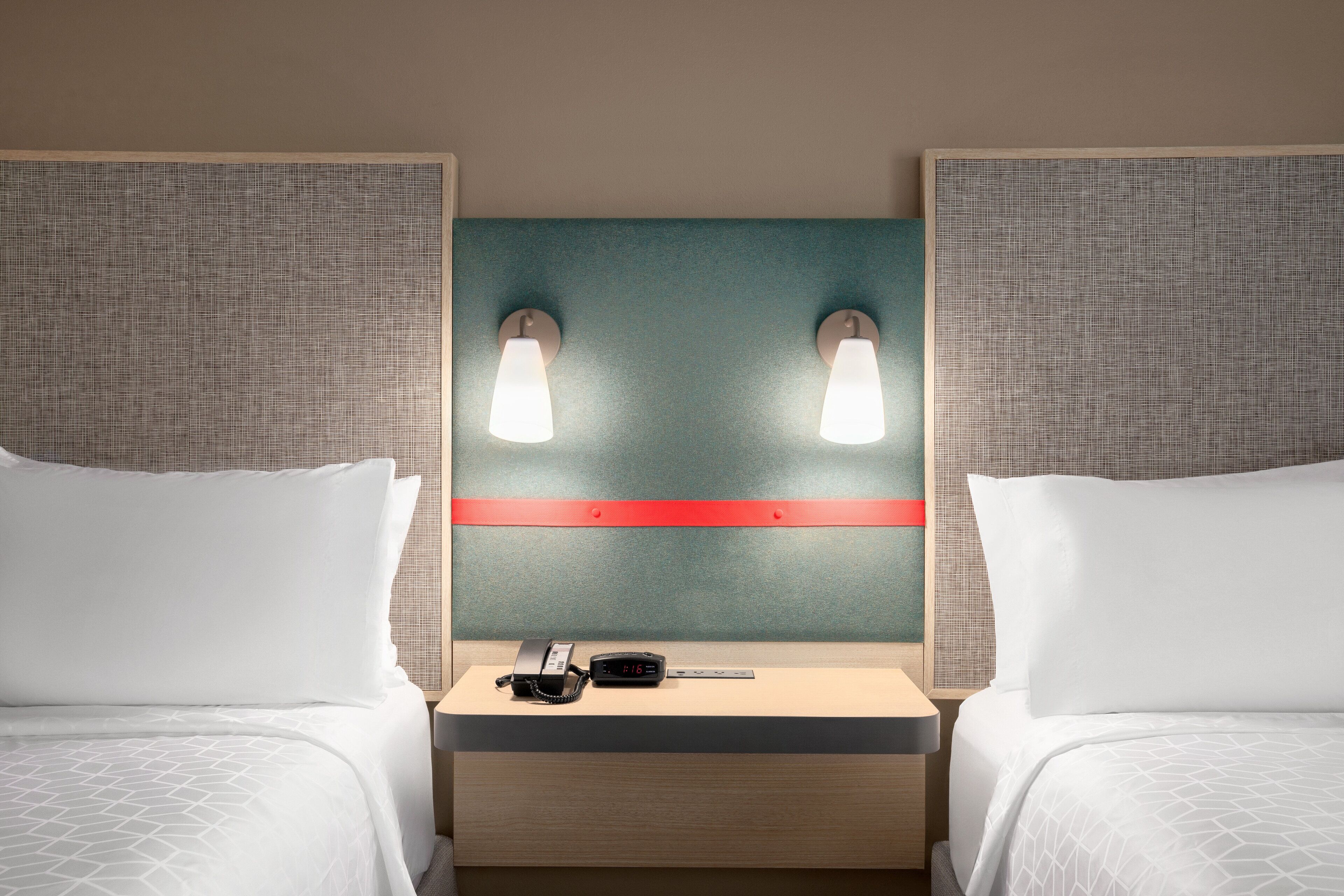 A hotel room with two beds, a nightstand with a phone and a clock, and wall-mounted lights.