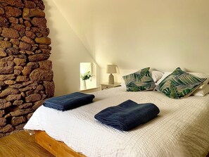 Ein Schlafzimmer mit Steinwand, einem Bett mit weißer Bettwäsche, zwei blauen Handtüchern und Kissen mit tropischem Muster.