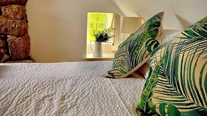 Une chambre avec un mur en pierre, une couette blanche matelassée et une vue sur la verdure par la fenêtre.