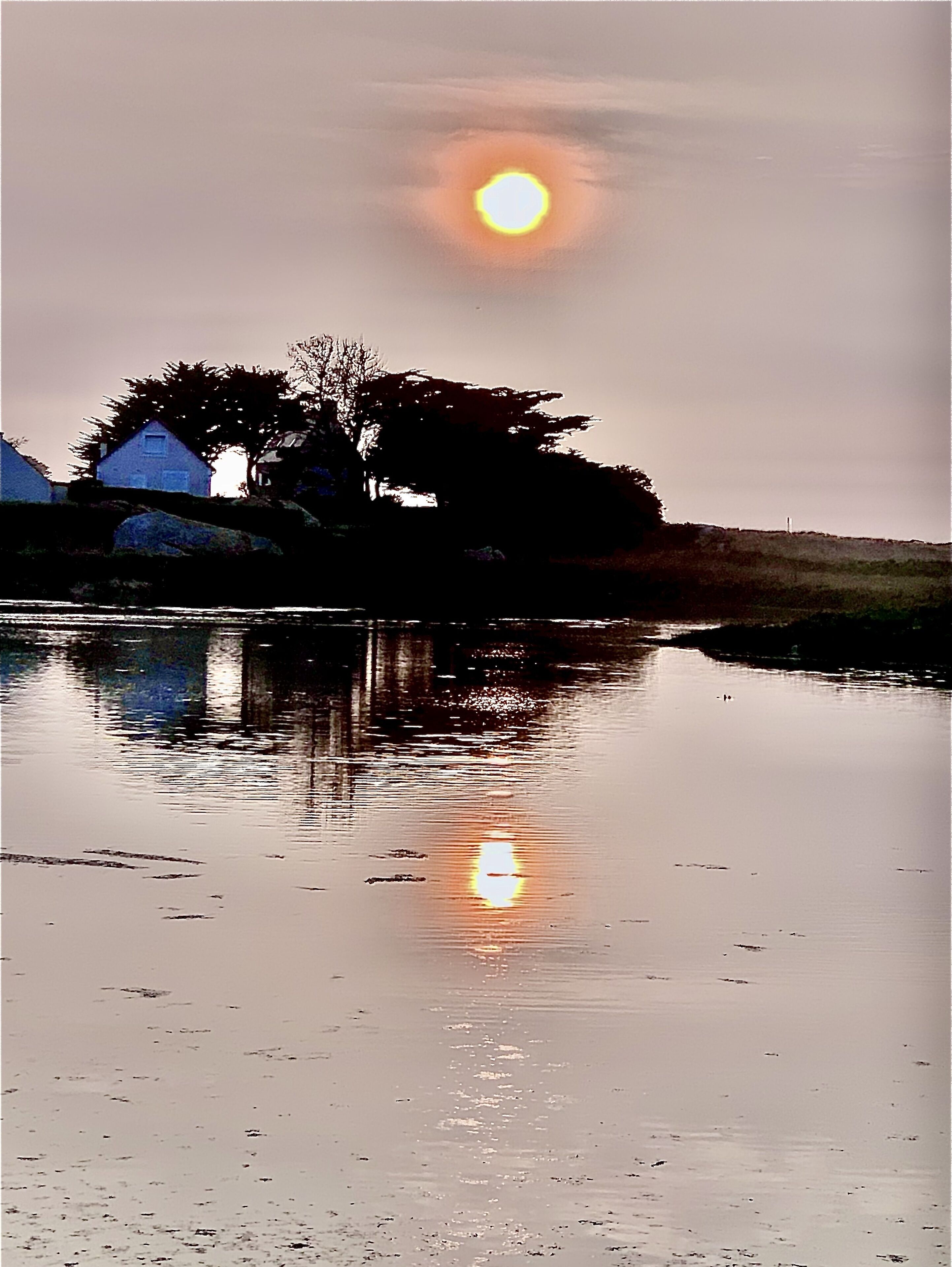A calm body of water reflecting the sun, with a silhouette of a house and trees in the background.