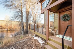 Eine Holzterrasse mit Geländer, die auf einen Fluss blickt, eine Steinstatue und eine Tür mit einem Kranz.