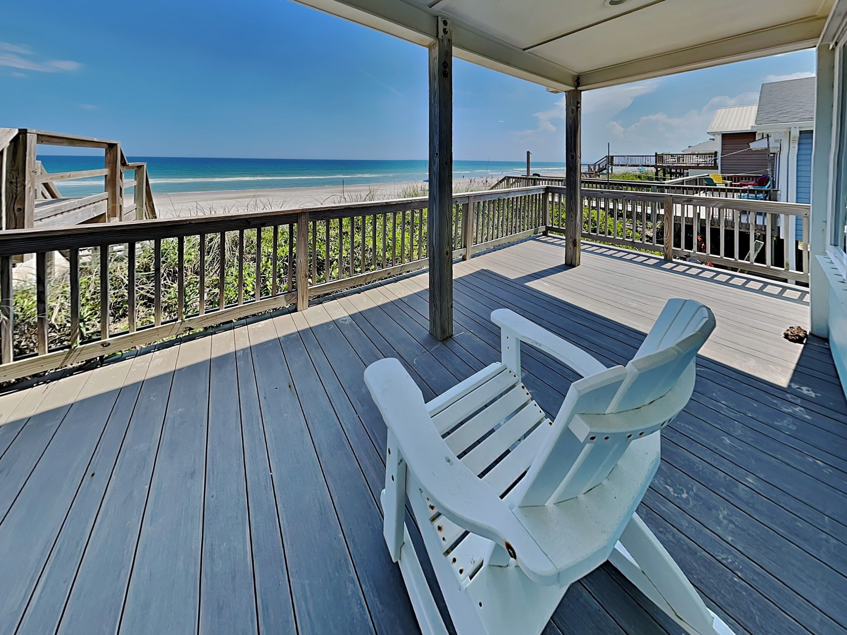 Eine Holzterrasse mit einem weißen Adirondack-Stuhl, mit Blick auf einen Strand mit einer Holzpromenade und Strandhäusern im Hintergrund.