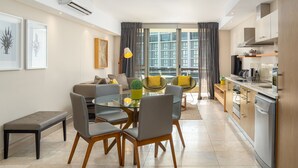 A modern living room with a glass dining table, grey chairs, a sofa, and a coffee table. There is a kitchen with a dishwasher, oven, and cabinets. The room has large windows with curtains and a view of buildings outside. - Spacious Studio Apartment on the Canals in CT (Cape Town)