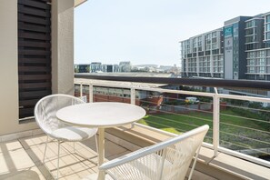 Un balcón con una mesa redonda y dos sillas blancas, con vistas a un paisaje urbano que incluye edificios modernos y un club náutico.