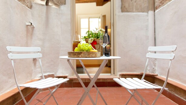 Un pequeño espacio al aire libre con dos sillas blancas y una mesa, que incluye una botella de vino, una copa y un frutero.