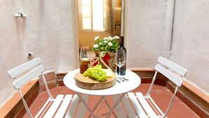 Una mesa redonda pequeña con una tabla de madera, dos sillas blancas, una botella de vino, una copa, un tarro de miel, un ramo de flores y un racimo de uvas.