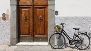 Unas puertas dobles de madera con un ventanal encima, una bicicleta con una cesta delantera y un aplique de pared a la derecha.