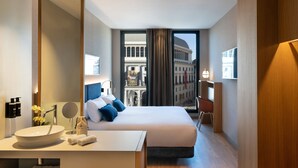 Une chambre d’hôtel avec un grand lit, une table de chevet avec un miroir, un lavabo rond et une vue sur un bâtiment historique par la fenêtre.