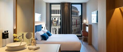A hotel room with a large bed, a bedside table with a mirror, a round sink, and a view of a historic building through the window.