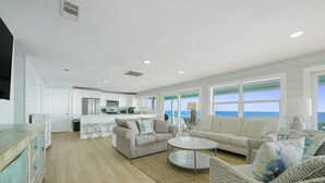 A spacious living area with a kitchen in the background, large windows offering a view of the ocean, and a cozy seating arrangement.