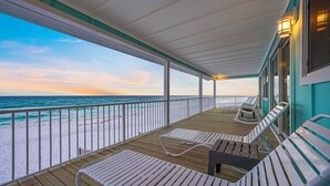 A covered deck with white cushioned chairs and a wooden table, overlooking a beach with a clear view of the ocean and a sunset sky.