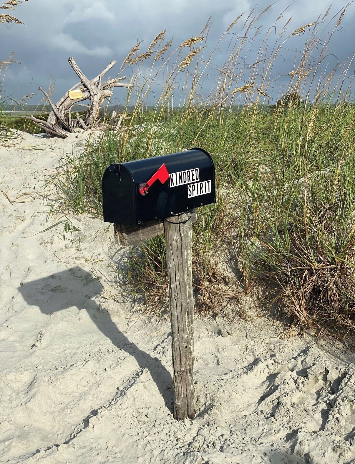 A mailbox with a red flag on a wooden post, surrounded by sand and grass.