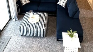 Un salon avec un canapé d’angle bleu foncé, un pouf rayé et une petite table blanche sur laquelle est posée une plante. Un tableau encadré est accroché au mur, au-dessus du canapé.